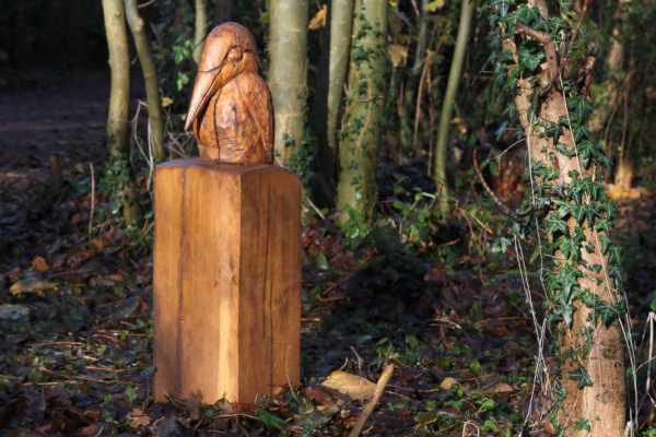 In front of some small trees is a polished carving of a large beaked bird possibly a Kingfisher atop a squat wooden post.