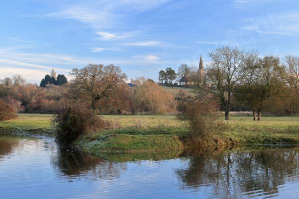 Against a bright blue sky a church with a steeple sits on a hill with grass and trees between it and the small lake in the foreground.