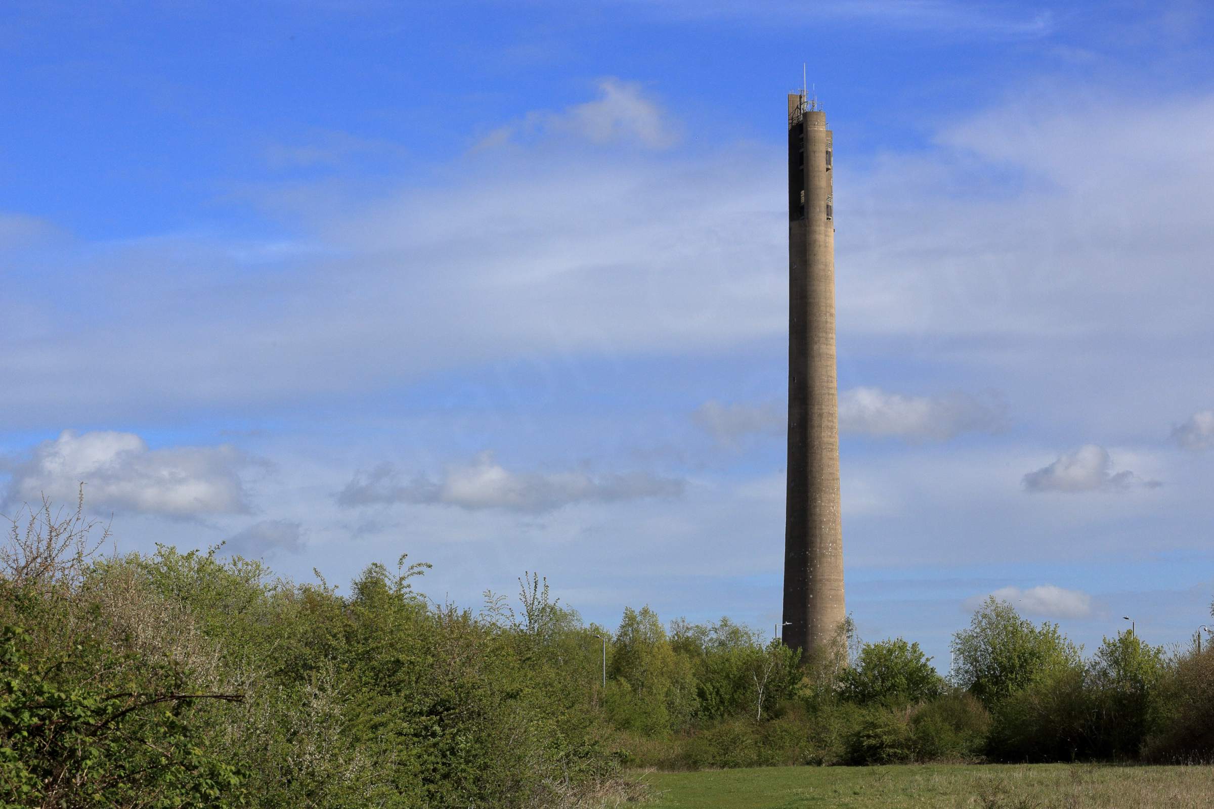 A tall slender lift tower dominates the skyline with a glimpse of meadow and hedgerow in the foreground.